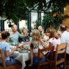 Familj samlad runt ett långbord med massa mat framdukat, sittandes i ett vackert uterum fyllt av gröna växter